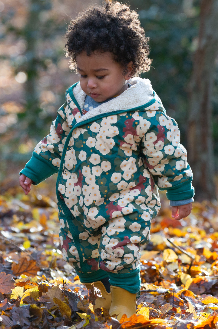 Baby walking in a forest wearing a sherpa fleece snowsuit with a green floral hedgerow print from Little Green Radicals - lined with organic cotton sherpa - to represent Little Green Radicals Autumn Winter clothing at Babipur.
