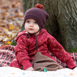 Baby sat on a blanket in the forest wearing a cosy chocolate brown knit hat with a crimson pom pom and chin straps, made from Fairtrade GOTS organic cotton by Little Green Radicals, shop now at Babipur.