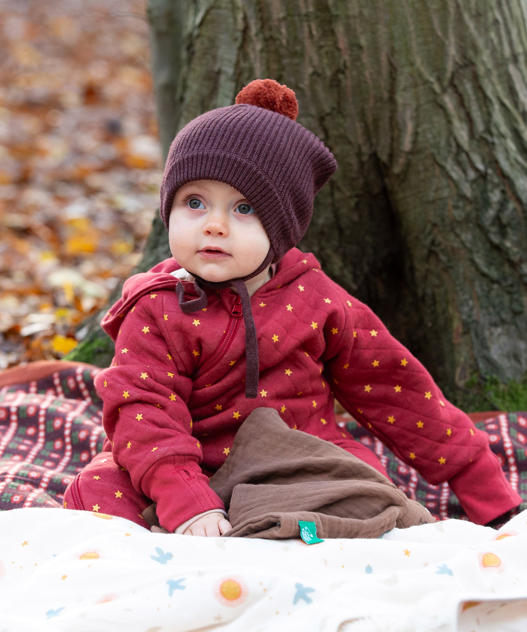 Baby sat on a blanket in the forest wearing a cosy chocolate brown knit hat with a crimson pom pom and chin straps, made from Fairtrade GOTS organic cotton by Little Green Radicals, shop now at Babipur.