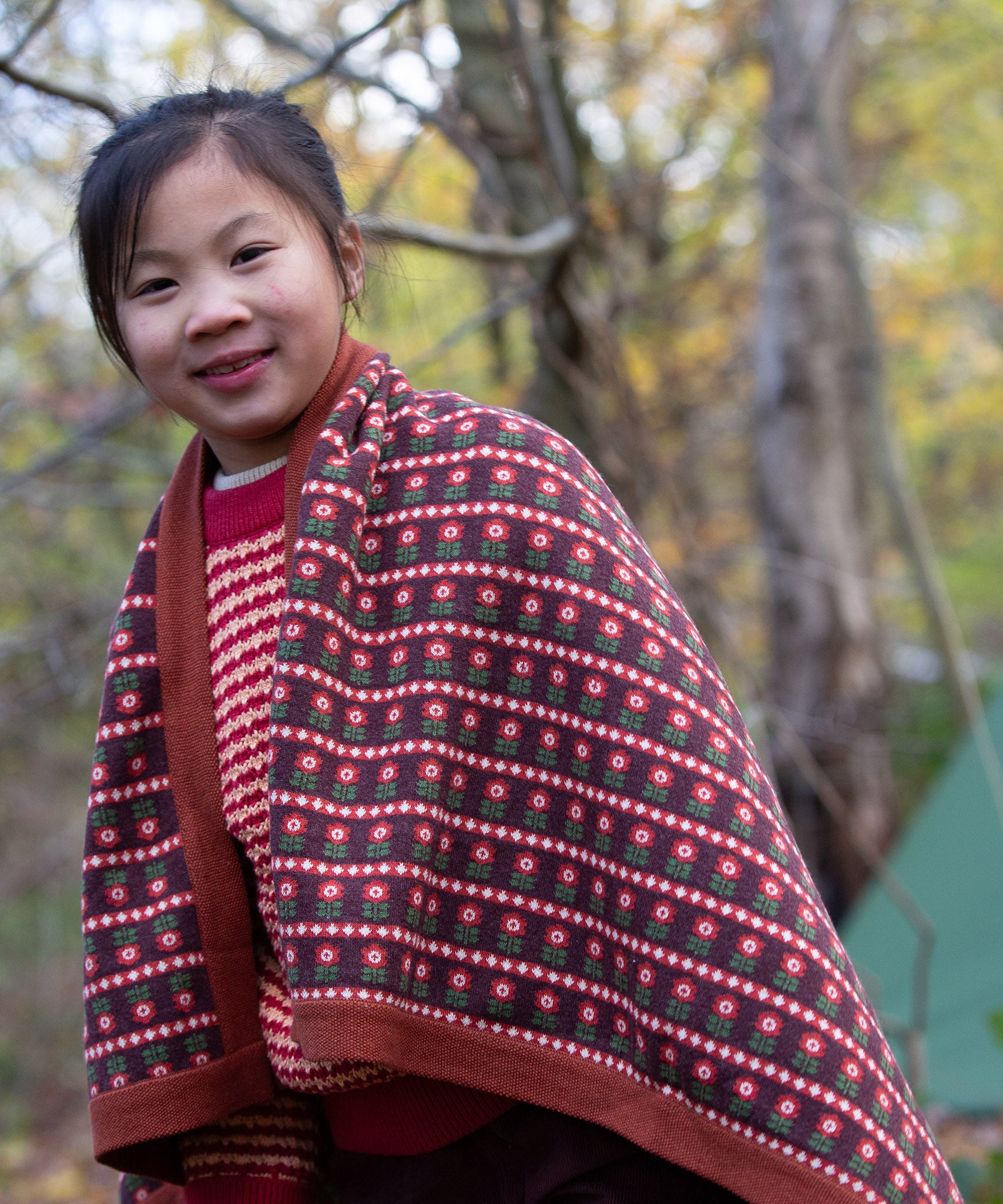 A knitted baby blanket with a brown and retro red floral pattern by Little Green Radicals — made from Fairtrade and GOTS-certified organic cotton — keeps a child cosy in the forest, available at Babipur.