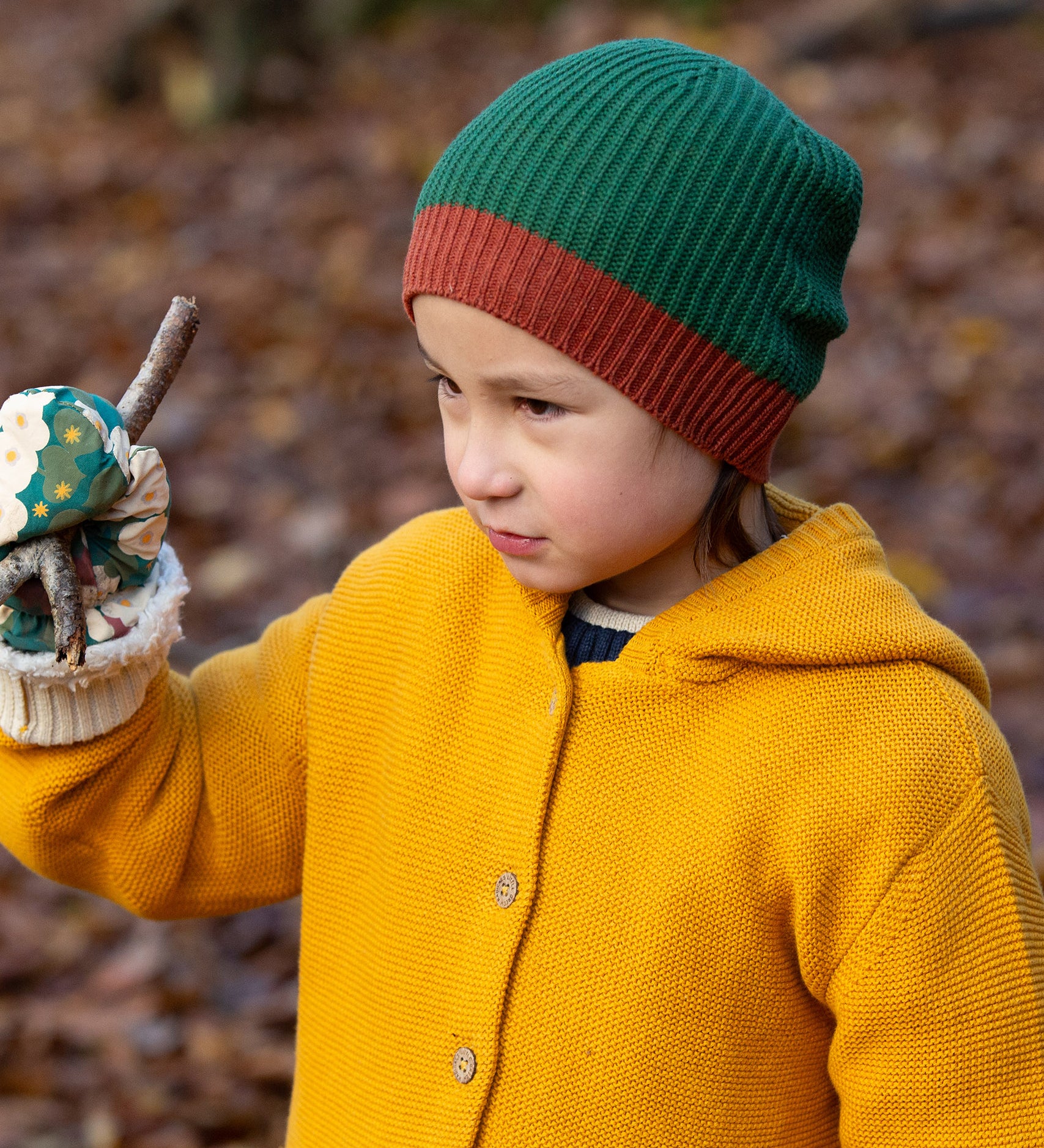 Close up of a child in a forest wearing a knitted beanie hat in chive green with a crimson made from soft Fairtrade and GOTS certified organic cotton by Little Green Radicals - for sale at Babipur.