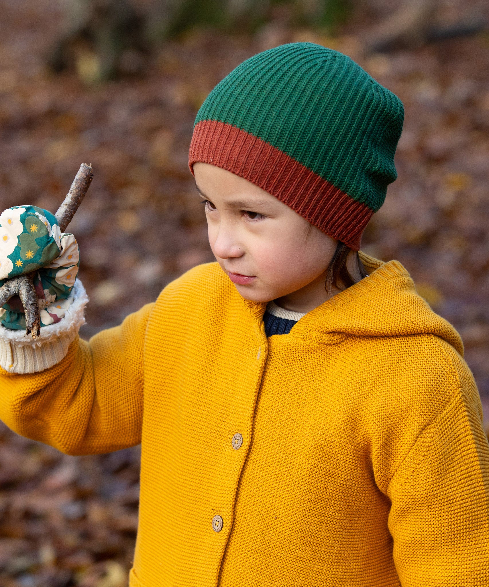 Close up of a child in a forest wearing a knitted beanie hat in chive green with a crimson made from soft Fairtrade and GOTS certified organic cotton by Little Green Radicals - for sale at Babipur.