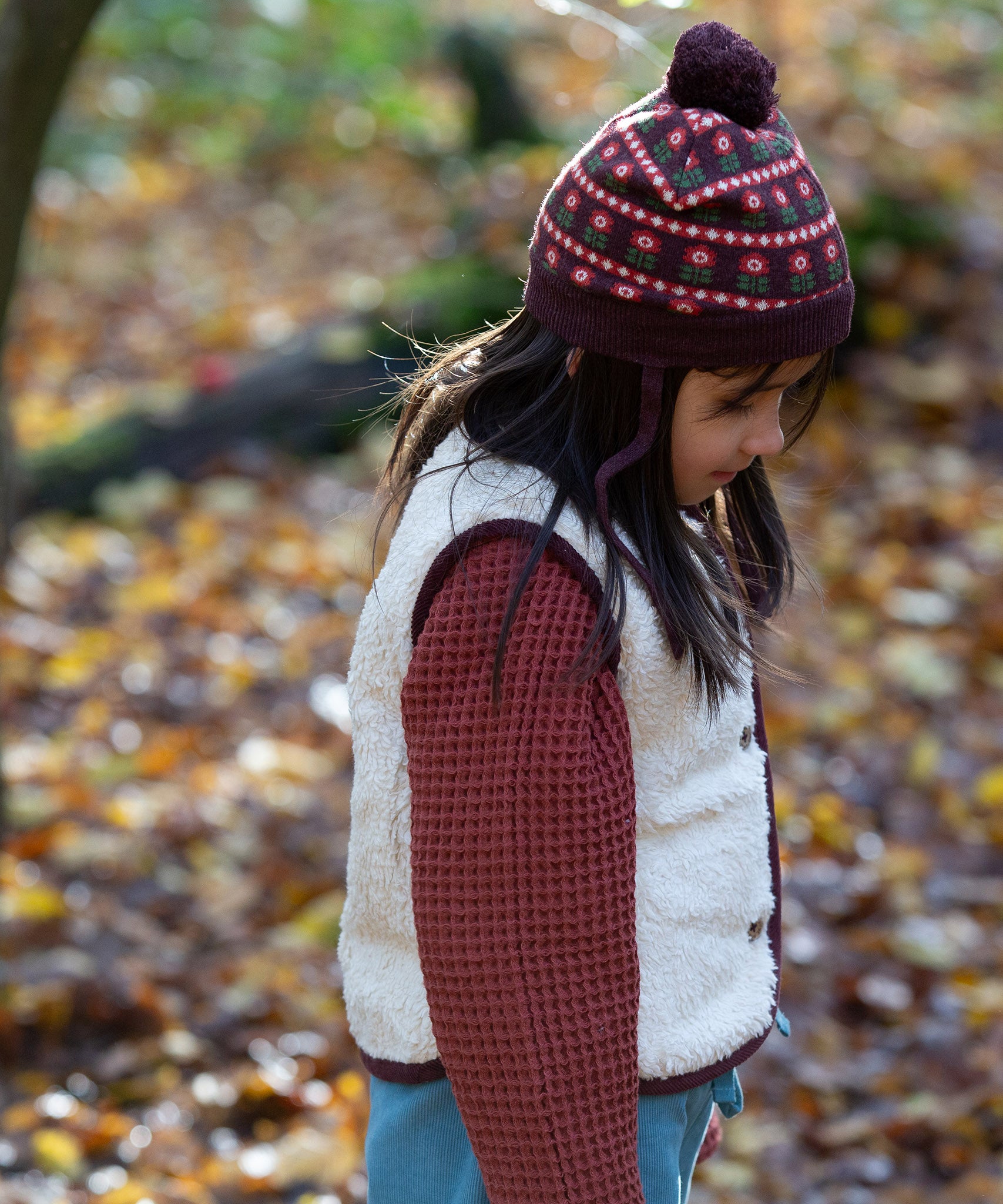 Child wearing an organic knit chocolate brown hat with a red floral pattern, brown pom pom and chin straps, by Little Green Radicals, available now at Babipur.