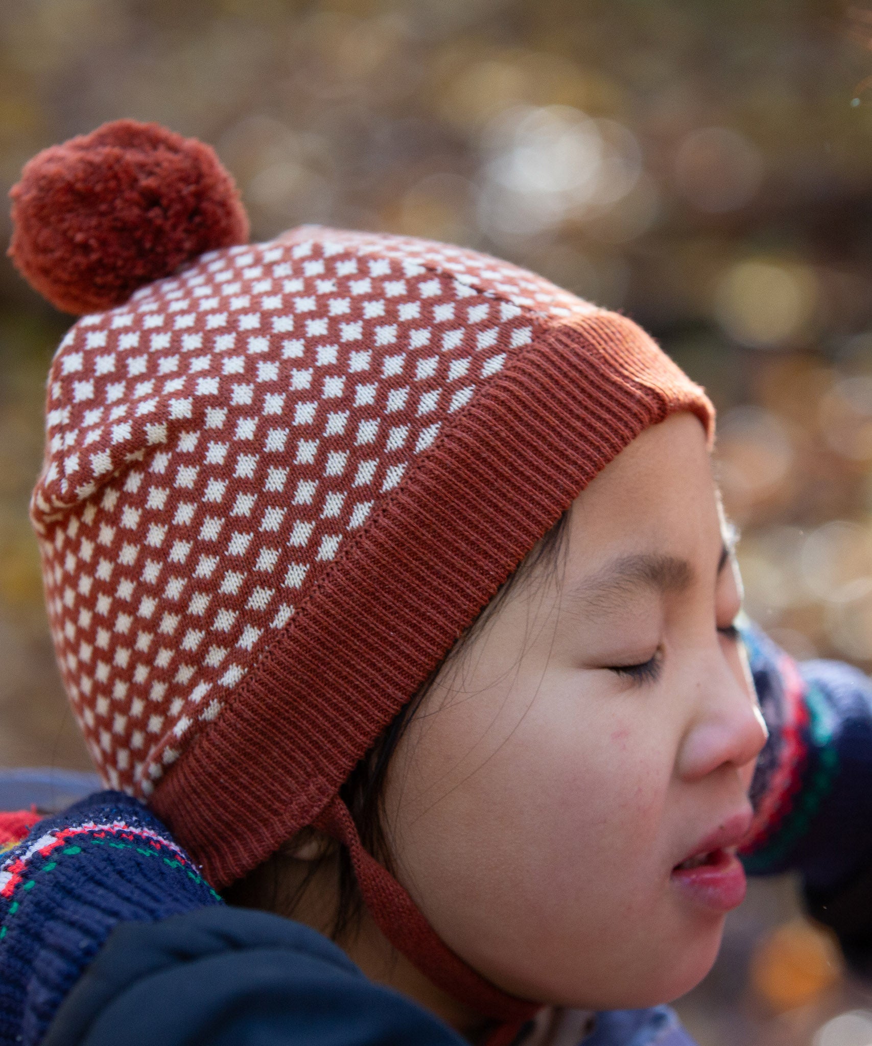 Close up of child wearing an organic cotton knit pom pom hat with ginger and cream diamon pattern, by Little Green Radicals, available to buy from Babipur. 