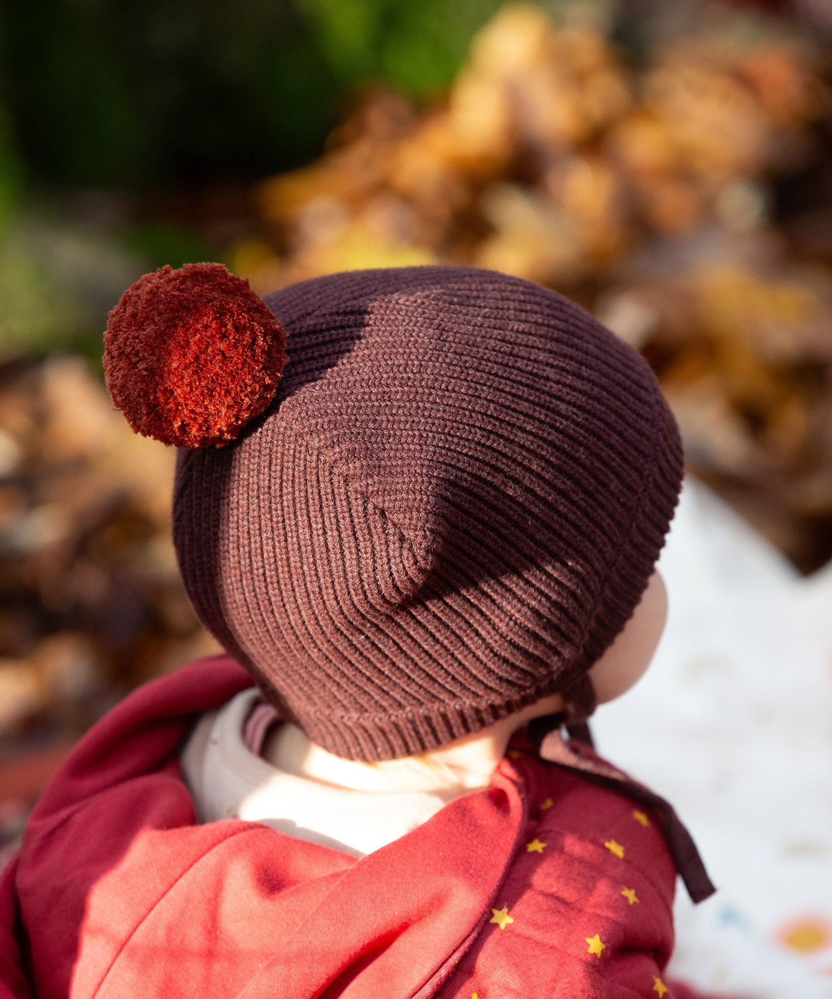 Back of baby's head wearing a Little Green Radicals knitted chocolate brown hat with a deep crimson pom pom, made from soft Fairtrade and GOTS certified organic cotton - available now at Babipur.