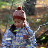 Front of child in a forest wearing a knitted ginger orange hat with a diamond pattern and a deep crimson pom pom, by Little Green Radicals, made from soft Fairtrade and GOTS certified organic cotton - shop now at Babipur.
