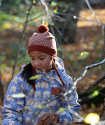 Front of child in a forest wearing a knitted ginger orange hat with a diamond pattern and a deep crimson pom pom, by Little Green Radicals, made from soft Fairtrade and GOTS certified organic cotton - shop now at Babipur.
