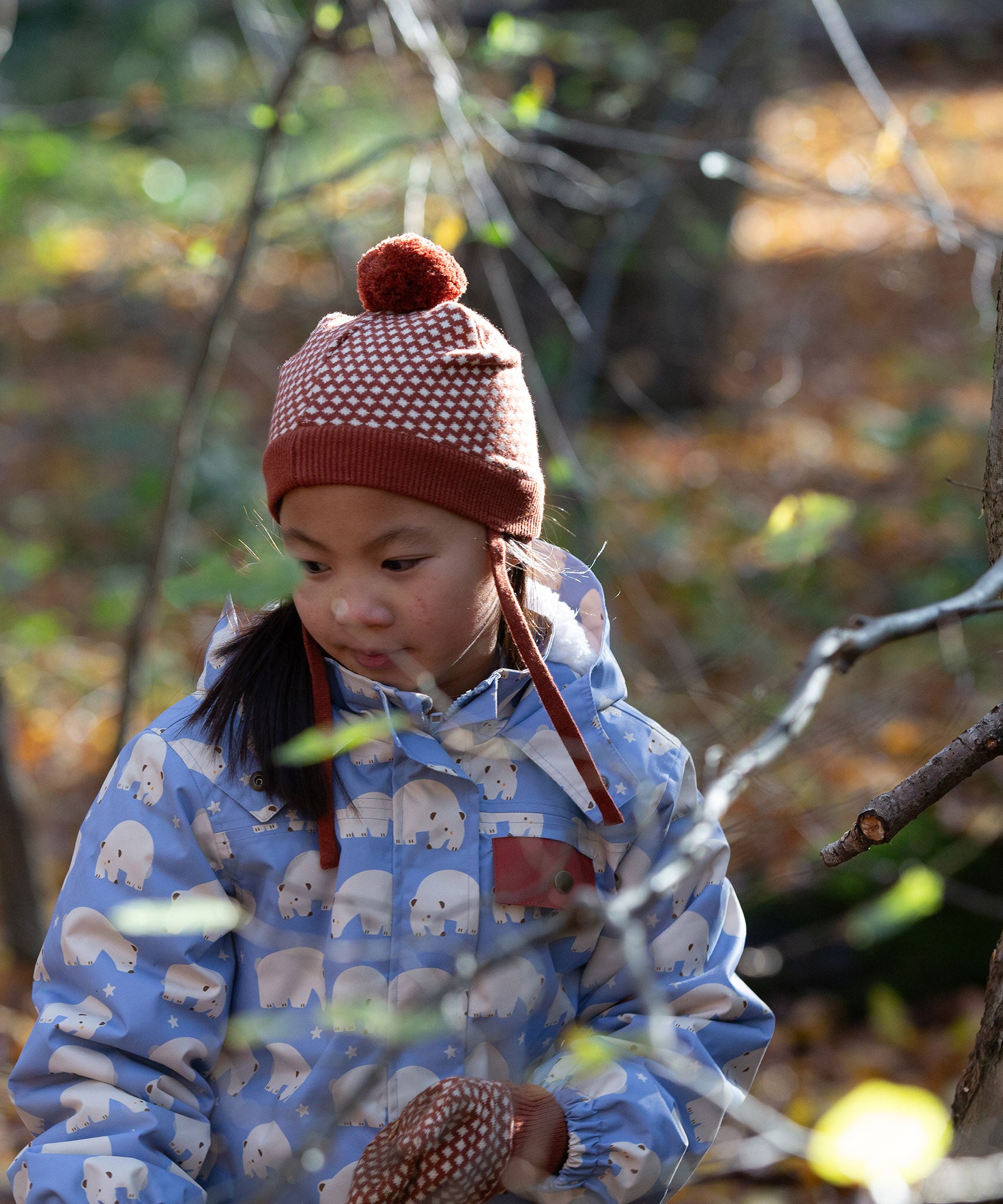 Front of child in a forest wearing a knitted ginger orange hat with a diamond pattern and a deep crimson pom pom, by Little Green Radicals, made from soft Fairtrade and GOTS certified organic cotton - shop now at Babipur.
