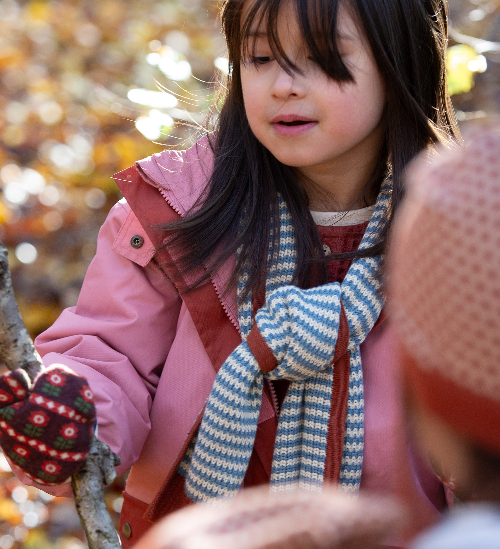 Blue and cream stripes adorn the knitted scarf worn by a child in the forest — designed by Little Green Radicals, produced from Fairtrade and GOTS-certified organic cotton, sold at Babipur.
