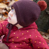 Close up of a baby in the forest wearing a knitted chocolate brown hat with a crimson pom pom, made from soft Fairtrade and GOTS certified organic cotton by Little Green Radicals - for sale at Babipur.