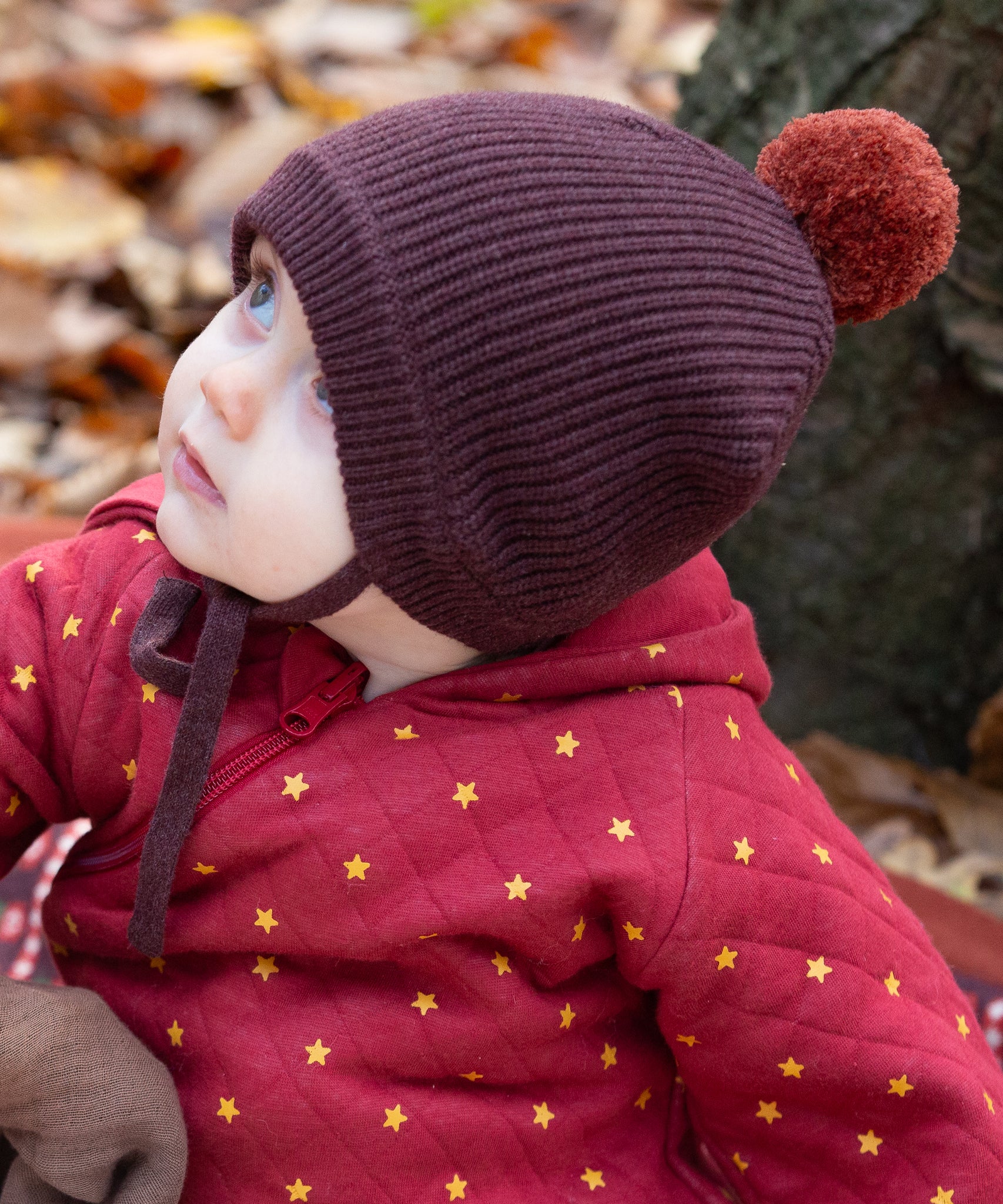 Close up of a baby in the forest wearing a knitted chocolate brown hat with a crimson pom pom, made from soft Fairtrade and GOTS certified organic cotton by Little Green Radicals - for sale at Babipur.