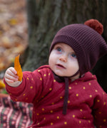 Close up of a baby holding a crispy autumn leaf wearing a knitted chocolate brown pom pom hat, made from soft Fairtrade and GOTS certified organic cotton by Little Green Radicals - available now at Babipur.
