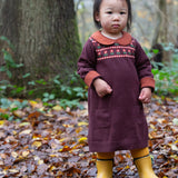 A child standing in woodland and wearing the dark maroon Little Green Radicals collecting flowers knitted collar dress.