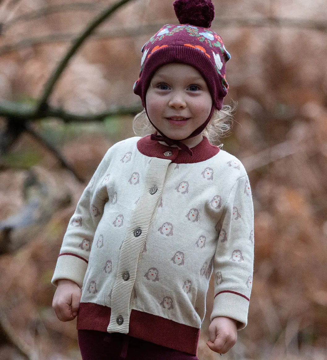 Little Green Radicals Fairtrade Organic Cotton Knitted Cardigan in Cream with Snow Penguins design and crimson cuffs, worn by child in the forest.