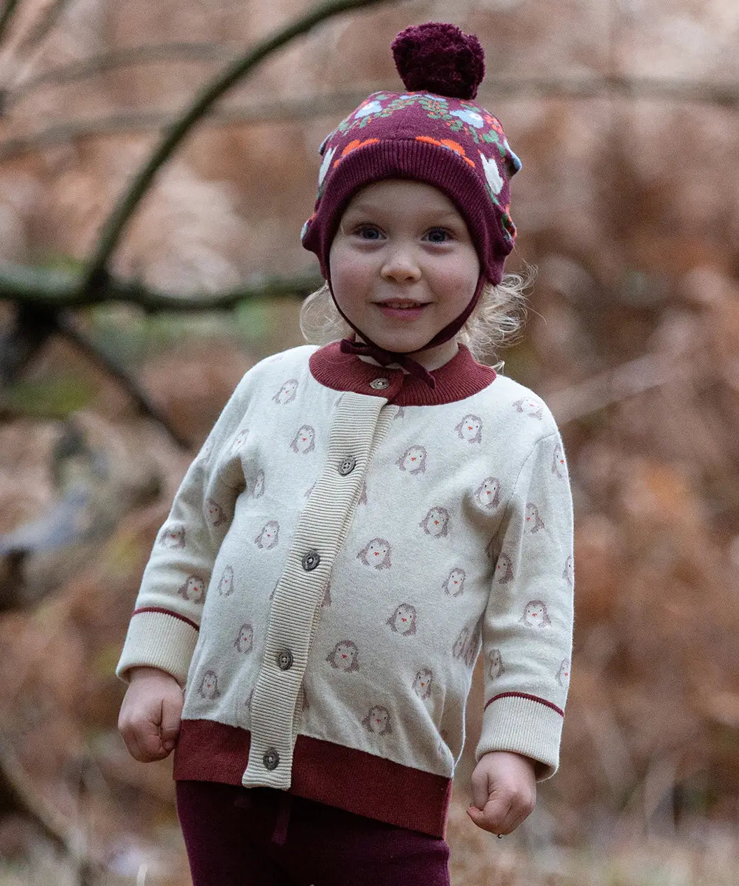 Little Green Radicals Fairtrade Organic Cotton Knitted Cardigan in Cream with Snow Penguins design and crimson cuffs, worn by child in the forest.