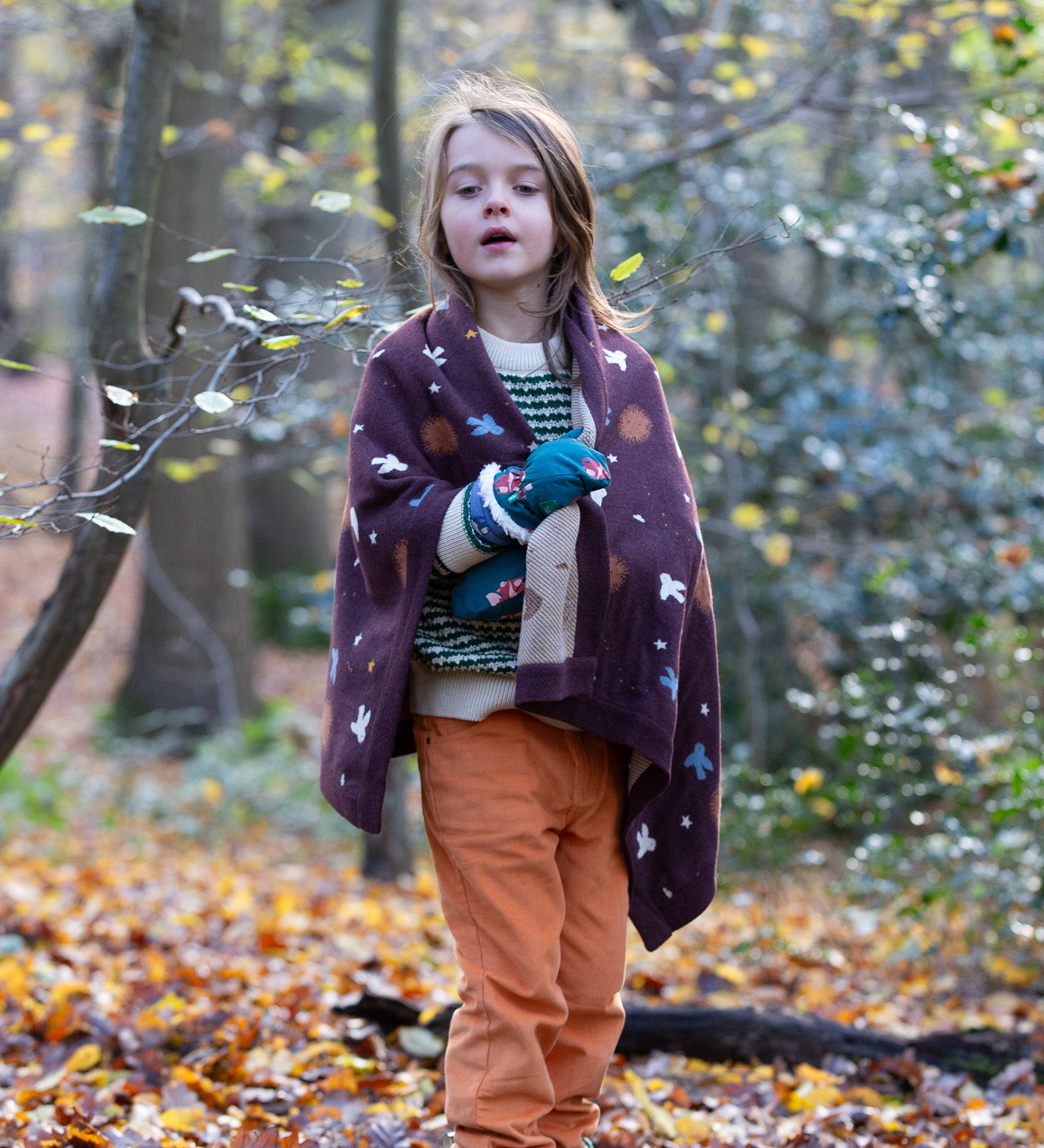 In the woods, a child is wrapped in a cosy brown baby blanket decorated with flying birds, designed by Little Green Radicals and made from Fairtrade and GOTS-certified organic cotton, for sale at Babipur.