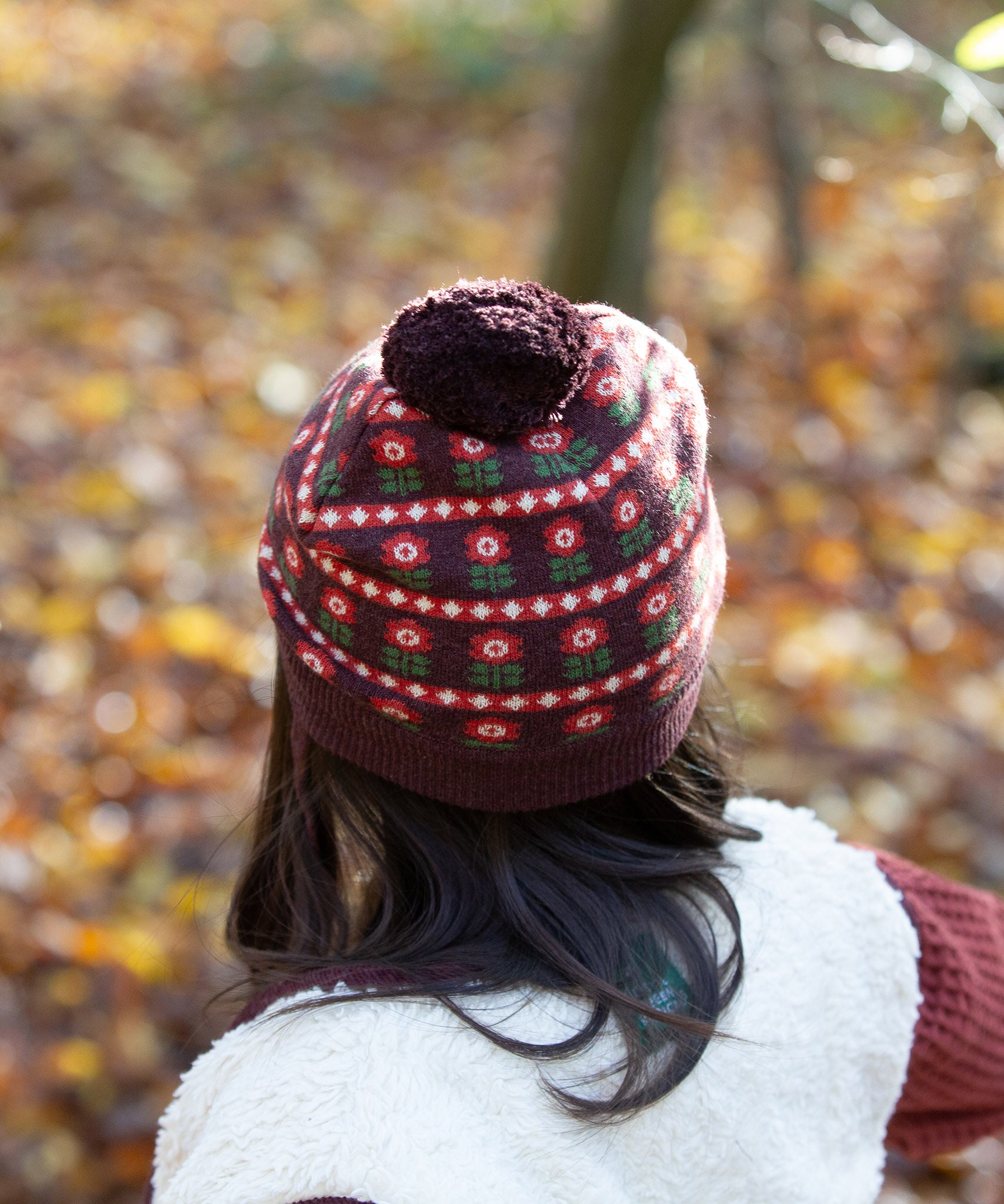 Back of child wearing an organic and Fairtrade knitted chocolate brown hat with a red floral pattern, brown pom pom, by Little Green Radicals, for sale at Babipur.