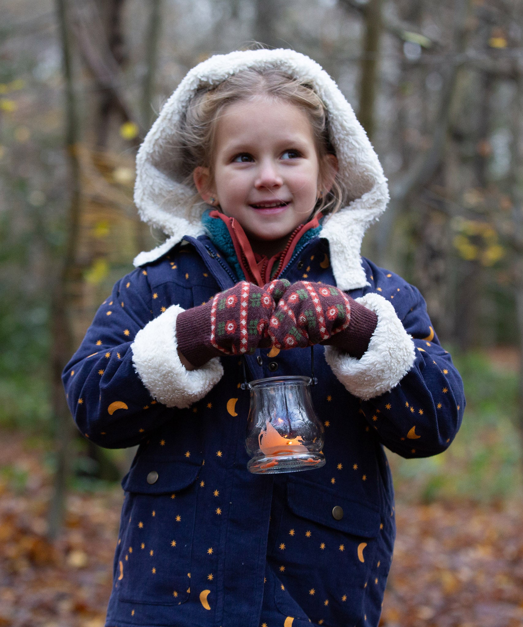 Child stood in a forest wearing brown and red floral Knitted Mittens by Little Green Radicals - made from soft GOTS organic and Fairtrade certified cotton - for sale at Babipur.
