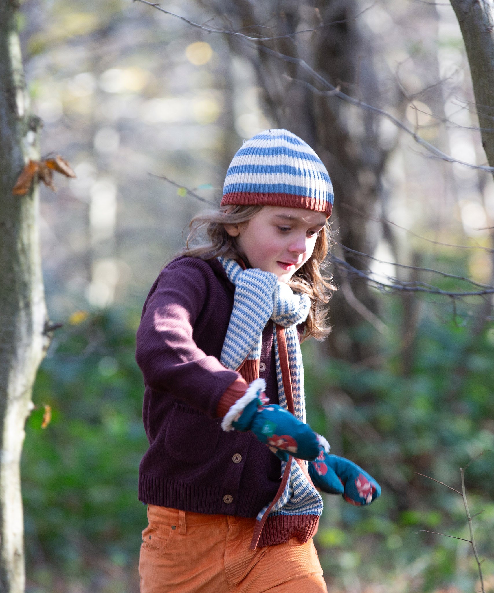 Child running through a forest wearing a striped blue and cream Knit Beanie by Little Green Radicals with a red trim and matching scarf - made from soft GOTS organic and Fairtrade certified cotton - available to buy at Babipur.
