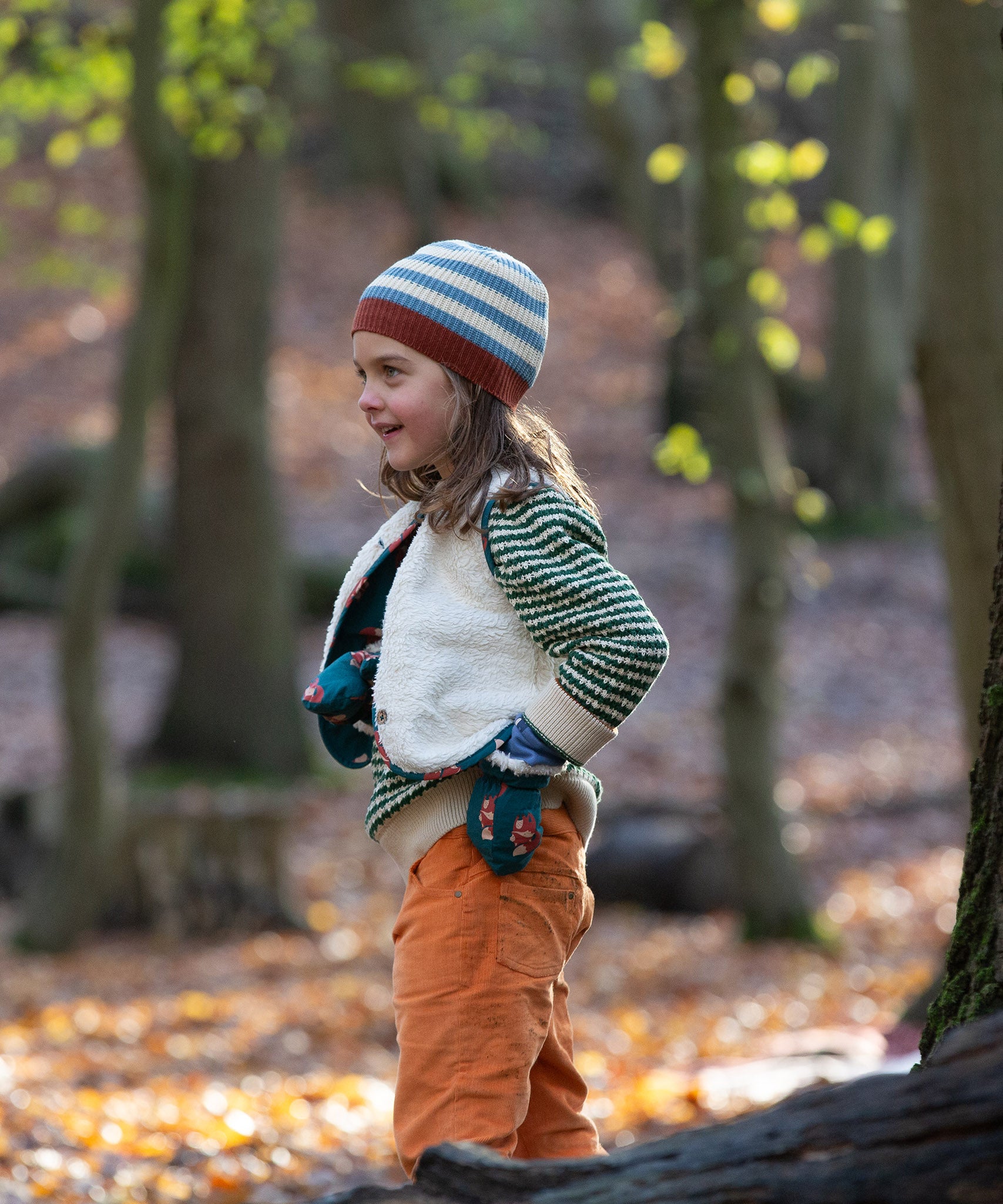 Child stood in a forest wearing a blue and cream striped Knitted Beanie by Little Green Radicals with a red trim - made from soft GOTS organic and Fairtrade certified cotton - for sale at Babipur.