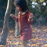 Child in a forest wearing Little Green Radicals Long-Sleeve Knitted Collar Dress in Ginger Orange - made from soft Fairtrade GOTS organic cotton - with Peter Pan Collar, striped cuffs, wooden buttons and two front pockets.