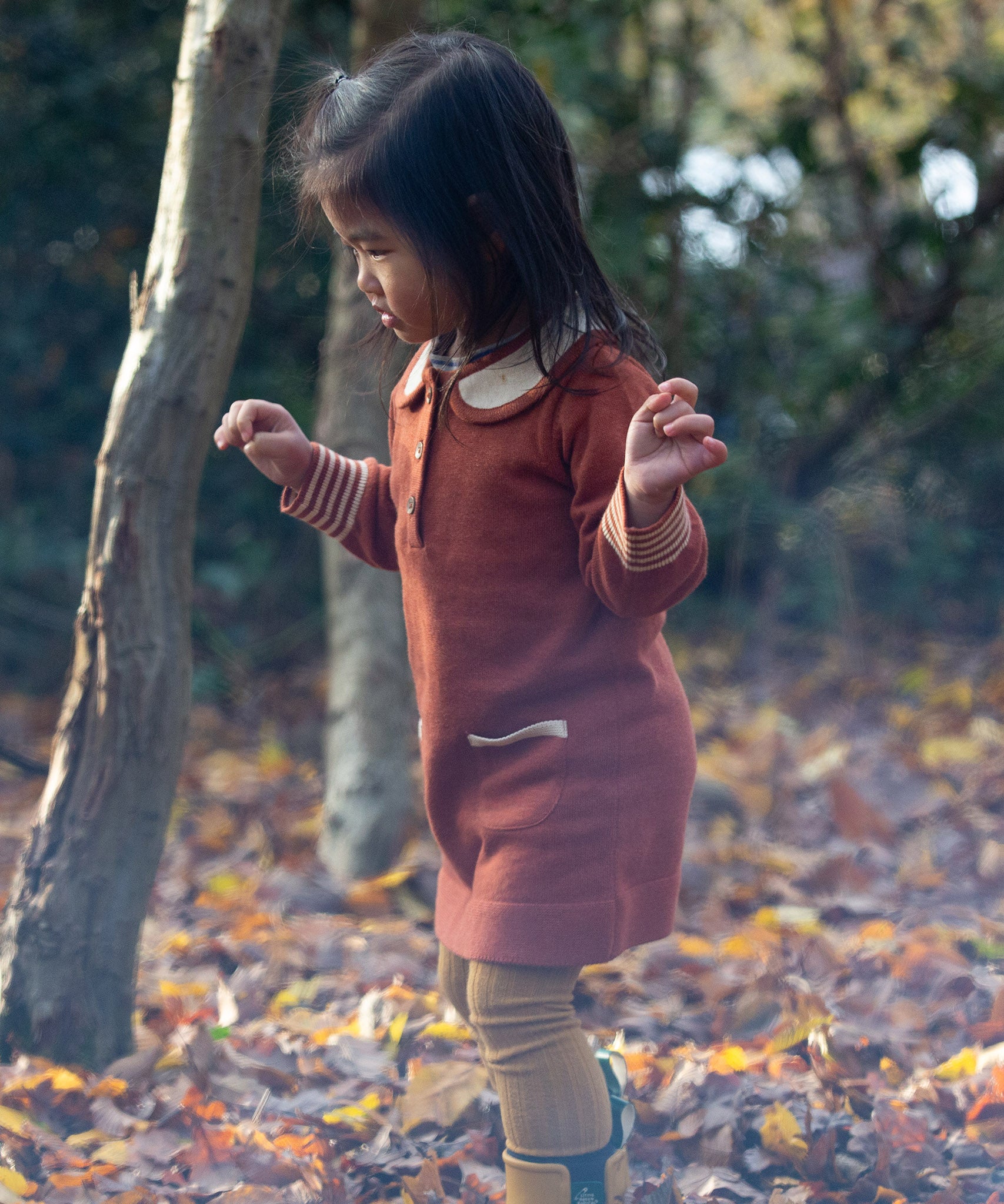 Child in a forest wearing Little Green Radicals Long-Sleeve Knitted Collar Dress in Ginger Orange - made from soft Fairtrade GOTS organic cotton - with Peter Pan Collar, striped cuffs, wooden buttons and two front pockets.