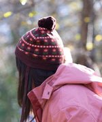 Side of child wearing a knitted brown hat with a red floral pattern and a brown pom pom, by Little Green Radicals, made from soft Fairtrade and GOTS certified organic cotton - shop now at Babipur.