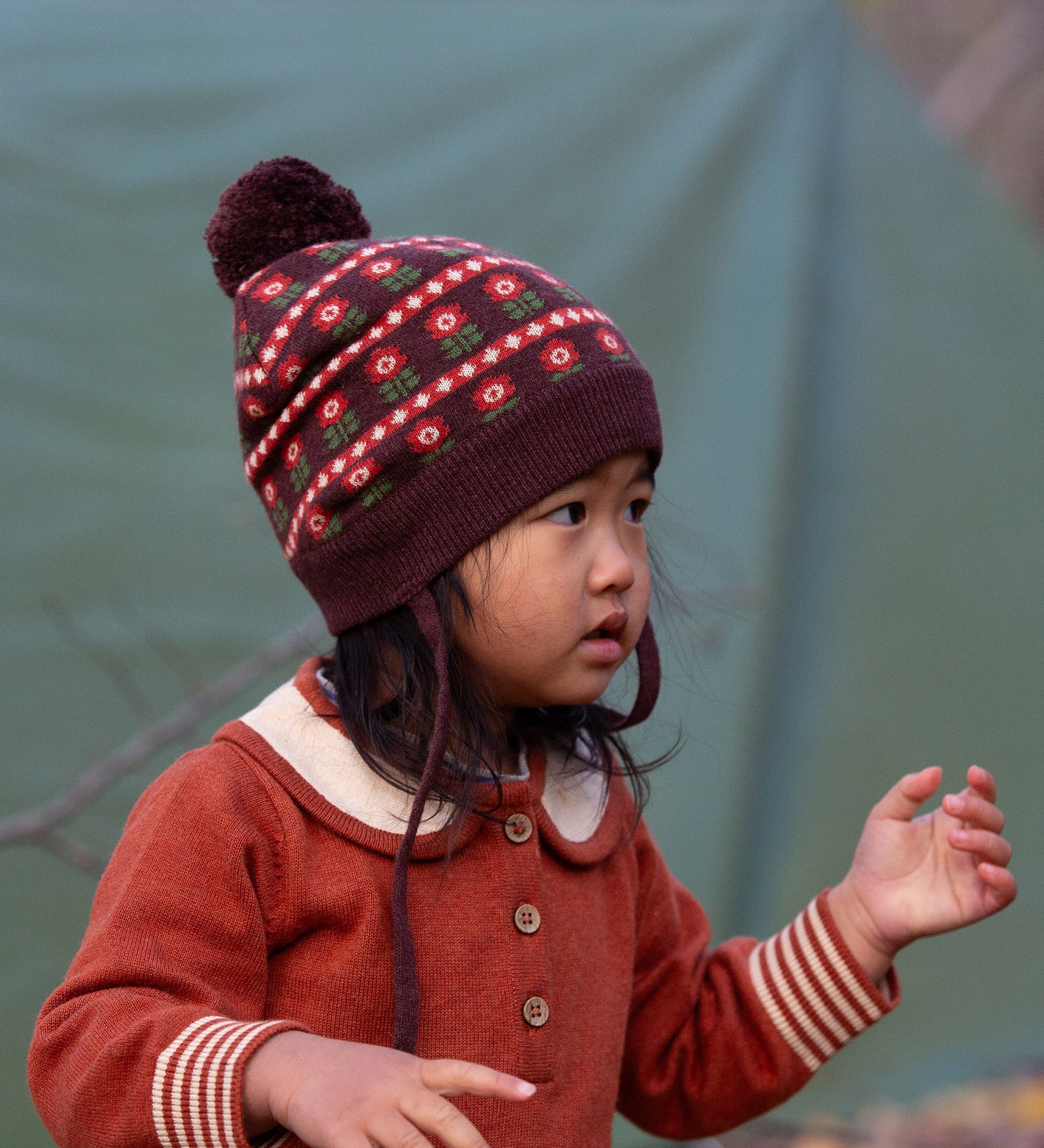 Close up of baby wearing a Little Green Radicals cosy chocolate brown knit hat with a floral pattern, brown pom pom and chin straps, made from Fairtrade GOTS organic cotton, available now at Babipur.