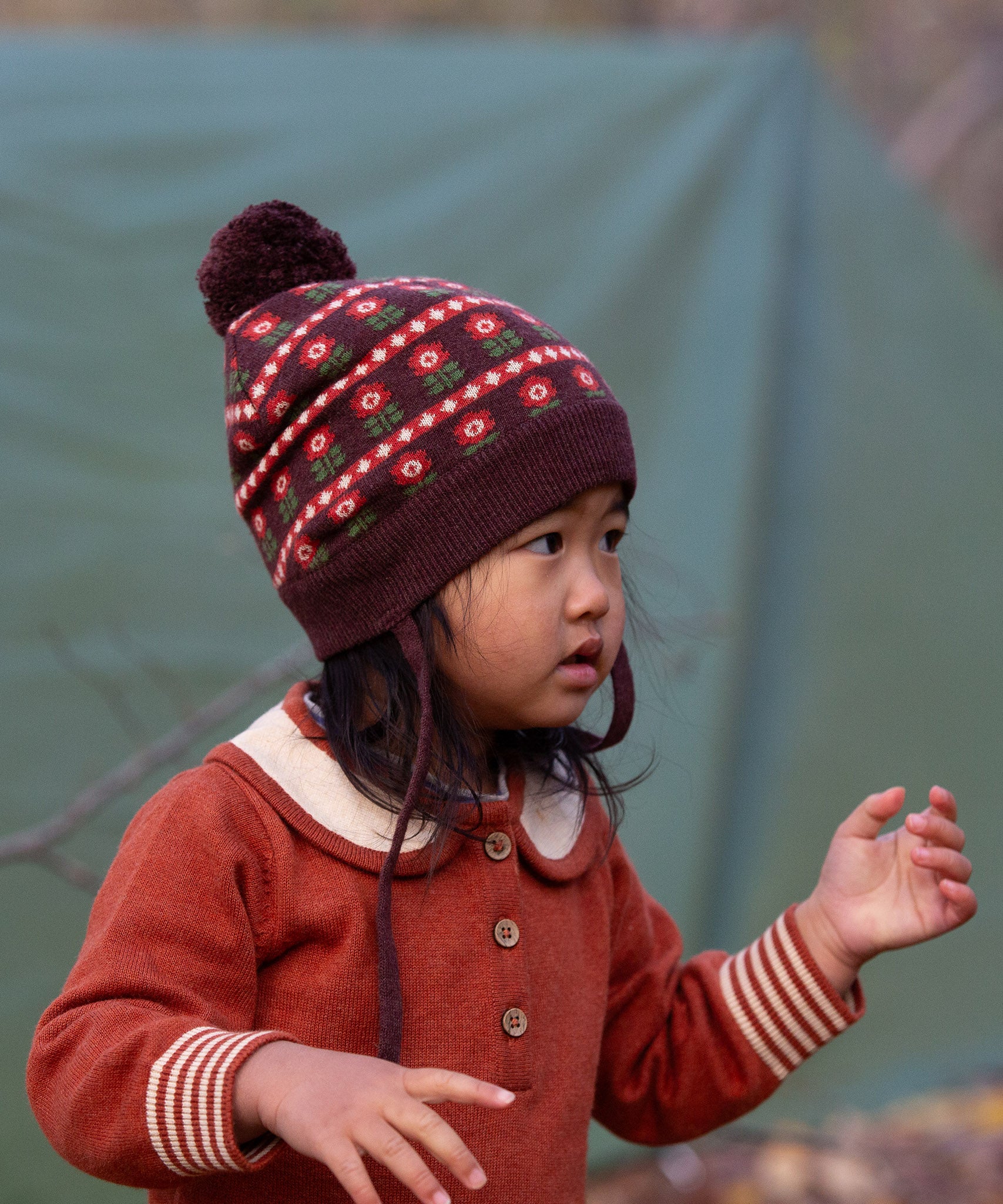 Close up of baby wearing a Little Green Radicals cosy chocolate brown knit hat with a floral pattern, brown pom pom and chin straps, made from Fairtrade GOTS organic cotton, available now at Babipur.