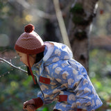 Side of child in a forest wearing a Little Green Radicals knitted ginger orange hat with a diamond pattern and a deep crimson pom pom, made from soft Fairtrade and GOTS certified organic cotton - available now at Babipur.