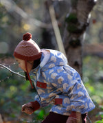 Side of child in a forest wearing a Little Green Radicals knitted ginger orange hat with a diamond pattern and a deep crimson pom pom, made from soft Fairtrade and GOTS certified organic cotton - available now at Babipur.