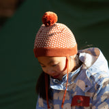 Child wearing an organic and Fairtrade Little Green Radicals knitted pom pom hat with an orange and cream pattern, for sale at Babipur.