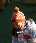 Child wearing an organic and Fairtrade Little Green Radicals knitted pom pom hat with an orange and cream pattern, for sale at Babipur.