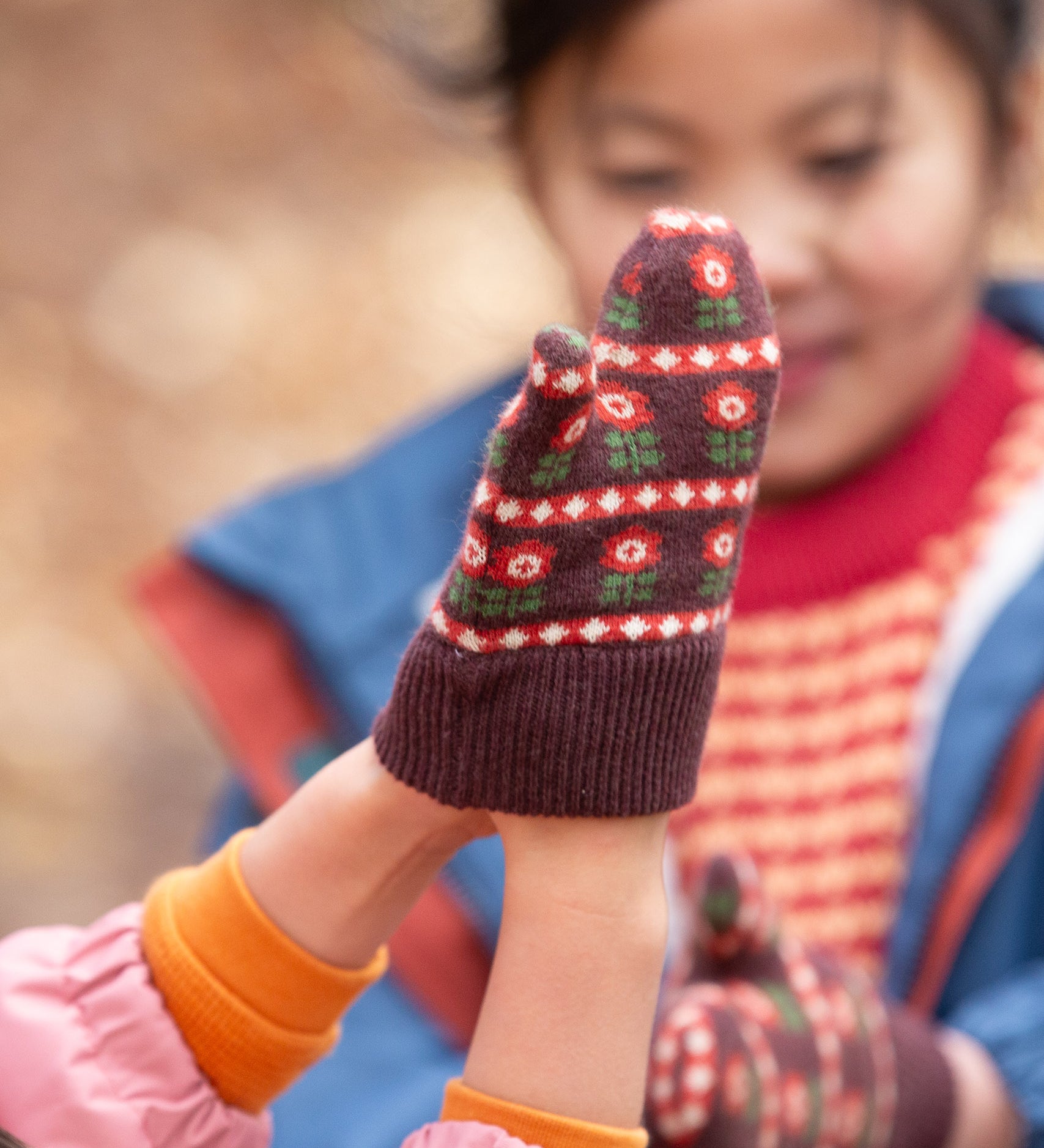 Close up of a child's hand wearing a knitted brown and red floral mitten from the Little Green Radicals Autumn Winter 2025 range - made from GOTS organic and Fairtrade certified cotton, available now at Babipur.