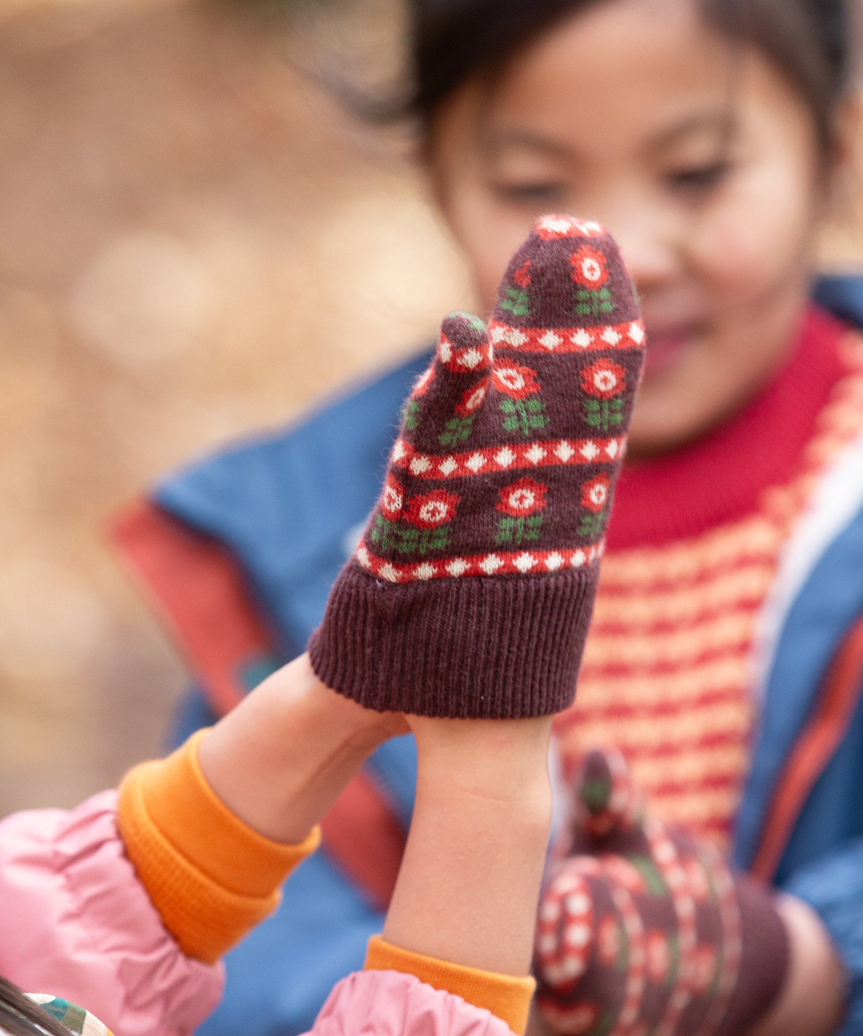 Close up of a child's hand wearing a knitted brown and red floral mitten from the Little Green Radicals Autumn Winter 2025 range - made from GOTS organic and Fairtrade certified cotton, available now at Babipur.
