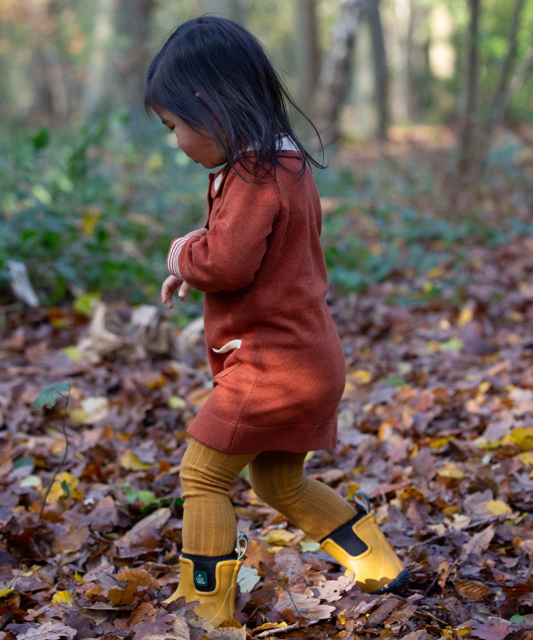 Child playing in a forest wearing a Long Sleeve Orange Ginger Knitte Dress by Little Green Radicals - made from soft GOTS organic and Fairtrade certified cotton - with striped cream and orange cuffs.
