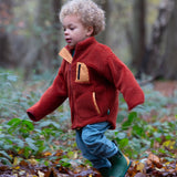 Boy in a forest wearing Little Green Radicals Polar Fleece Jacket in Ginger red with yellow zipped pockets, made from recycled fleece, for sale at Babipur.
