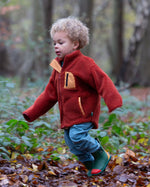 Boy in a forest wearing Little Green Radicals Polar Fleece Jacket in Ginger red with yellow zipped pockets, made from recycled fleece, for sale at Babipur.
