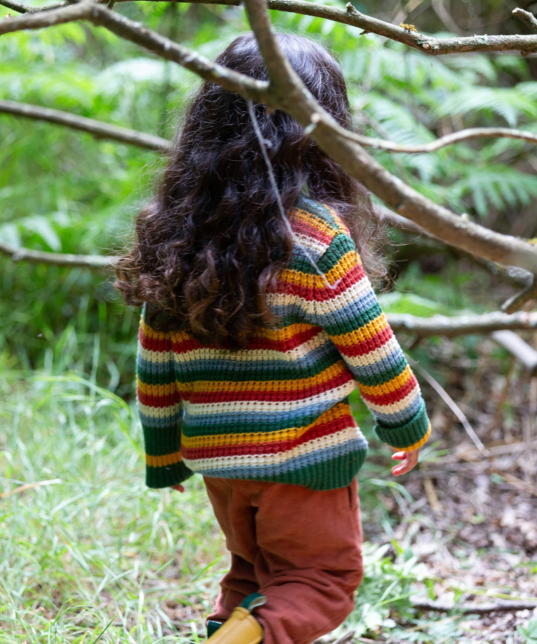 A child walking through woodland and wearing the Little Green Radicals rainbow waffle snuggly knitted jumper