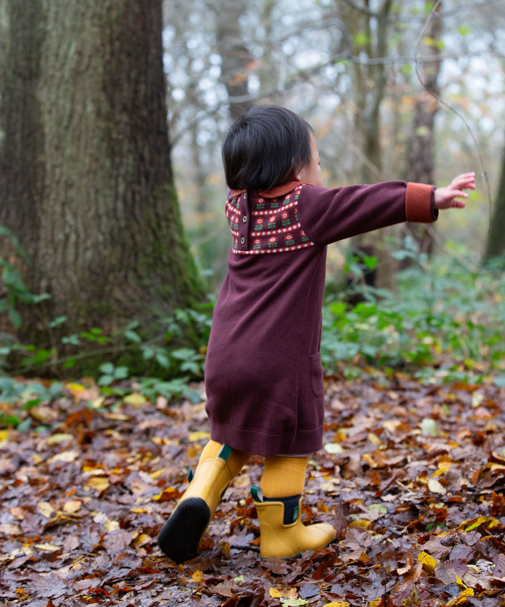 A child playing in woodland and wearing the dark maroon Little Green Radicals collecting flowers knitted collar dress.