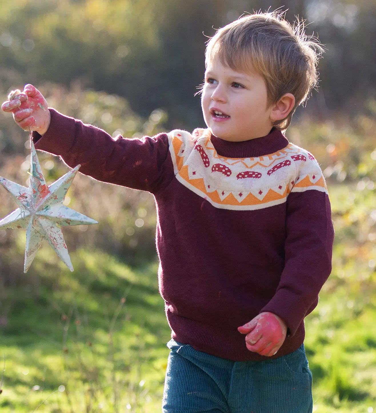 Little Green Radicals Knitted Organic Cotton Jumper in burgundy with Mushroom Fair Isle design worn by child in the forest.