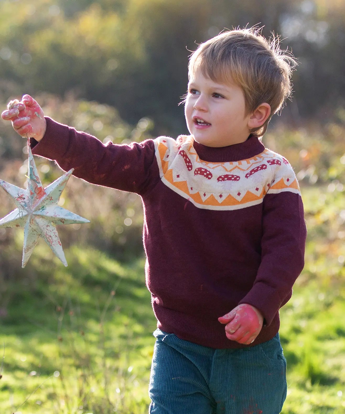 Little Green Radicals Knitted Organic Cotton Jumper in burgundy with Mushroom Fair Isle design worn by child in the forest.