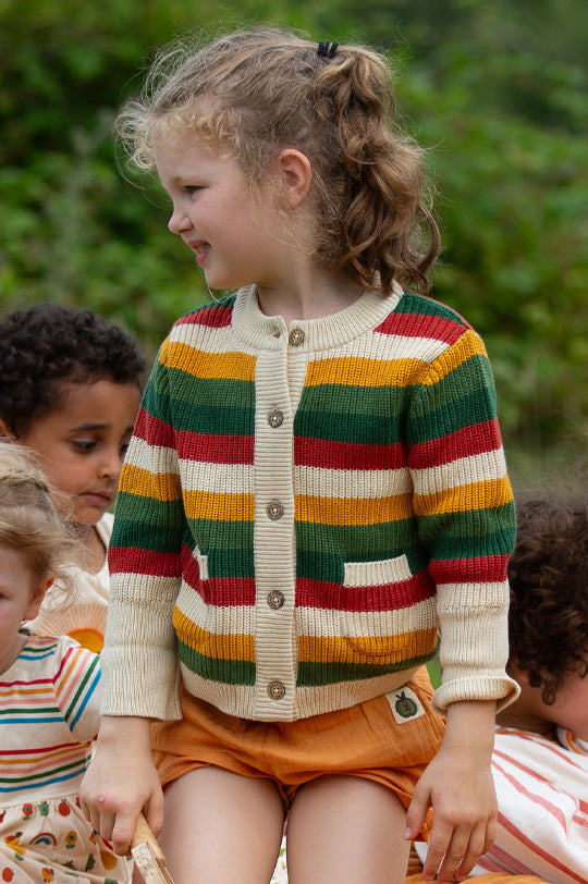 Child outdoors wearing Little Green Radicals Knitted Snuggly Cardigan in thick rainbow stripes with wooden buttons, made from organic cotton, paired with muslin yellow shorts.