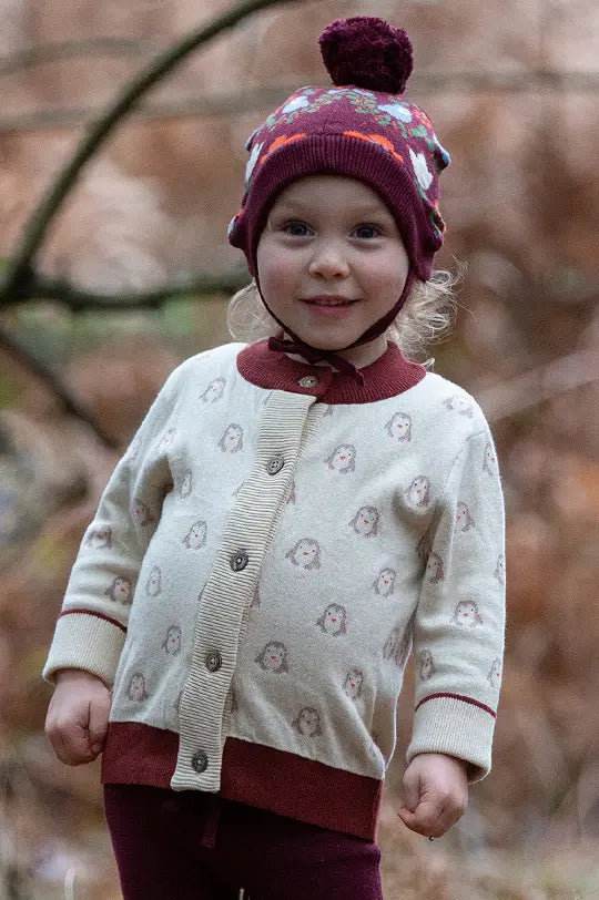 Little Green Radicals knitted cardigan in white penguin design with crimson neckline and wooden buttons, worn by child in a forest wearing a knitted bobble hat in plum by Little Green Radicals.