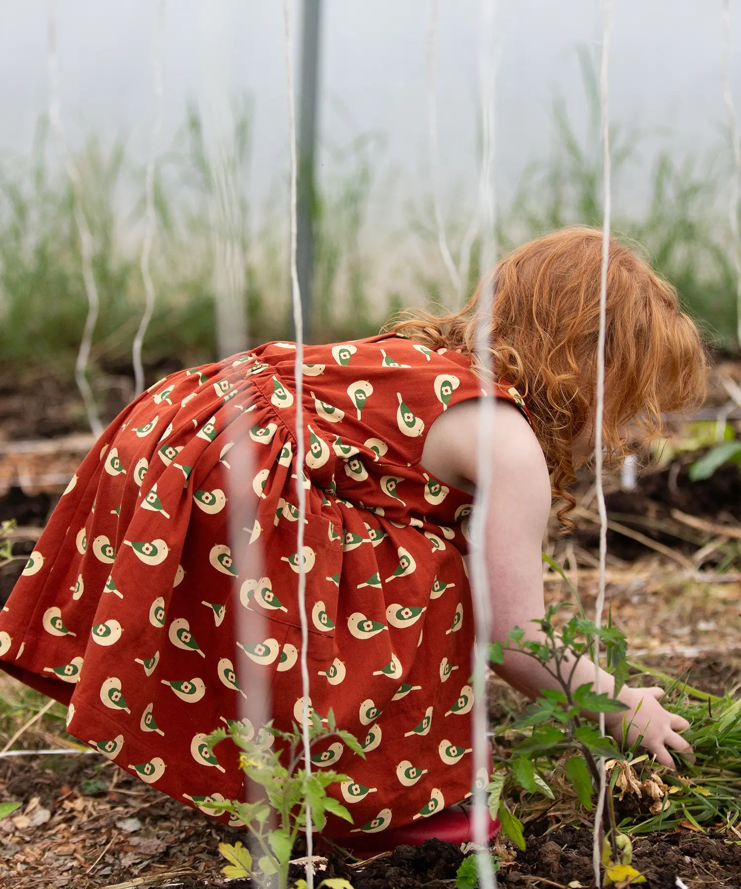 Little Green Radicals red spring birds print collared pinafore dress on a child gardening.