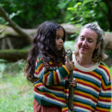A close up of a child standing in the woods and wearing the Little Green Radicals rainbow waffle snuggly knitted jumper with an adult in the background wearing a matching rainbow stripe jumper