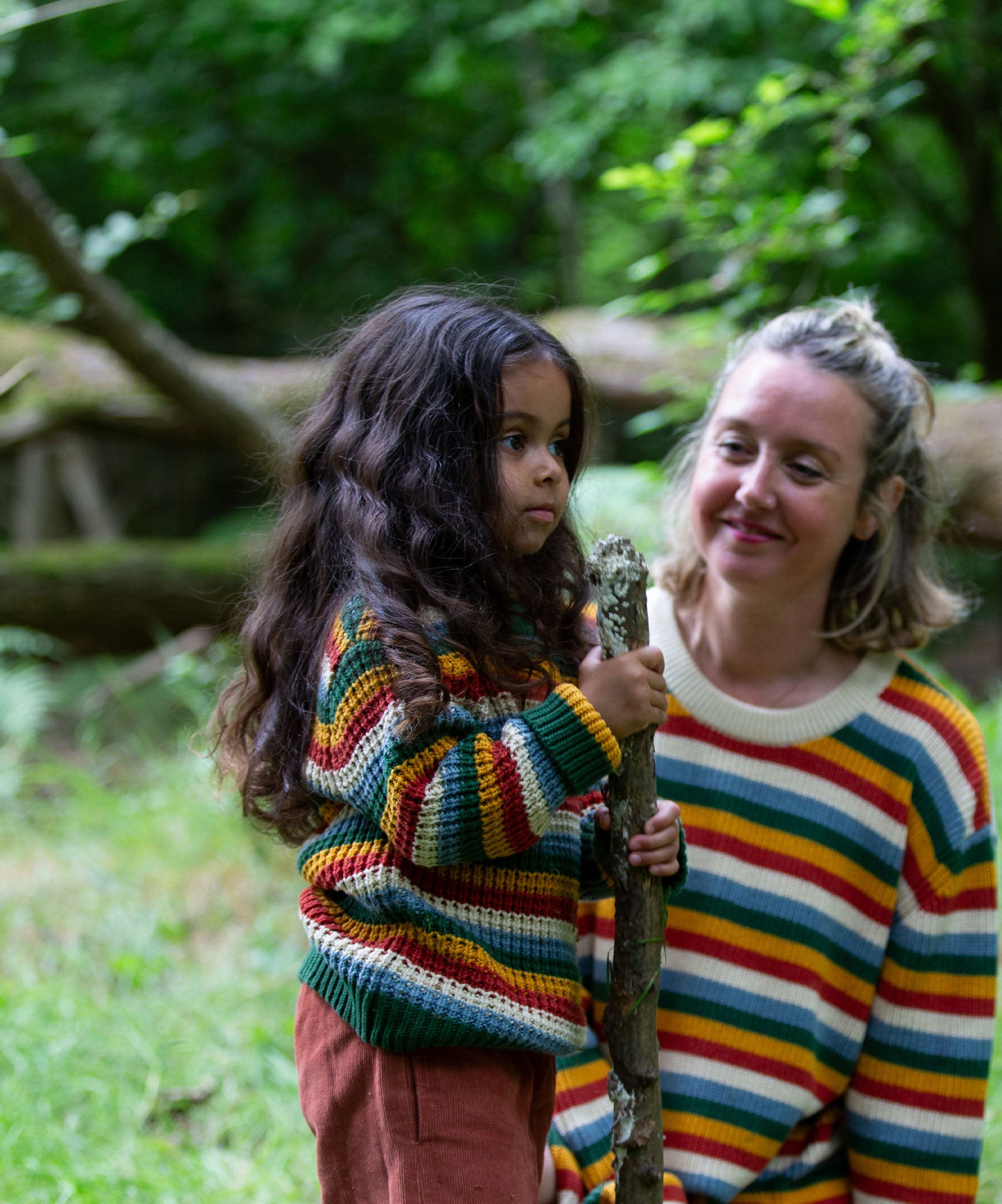 A close up of a child standing in the woods and wearing the Little Green Radicals rainbow waffle snuggly knitted jumper with an adult in the background wearing a matching rainbow stripe jumper