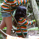 A close up of a child playing in the woods and wearing the Little Green Radicals rainbow waffle snuggly knitted jumper with an adult wearing a matching jumper