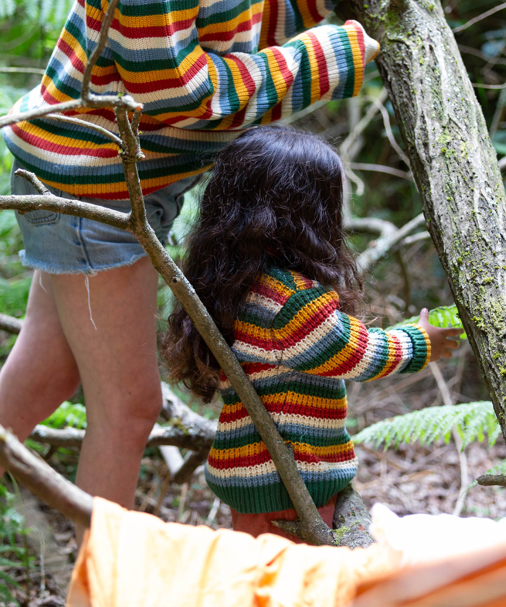 A close up of a child playing in the woods and wearing the Little Green Radicals rainbow waffle snuggly knitted jumper with an adult wearing a matching jumper