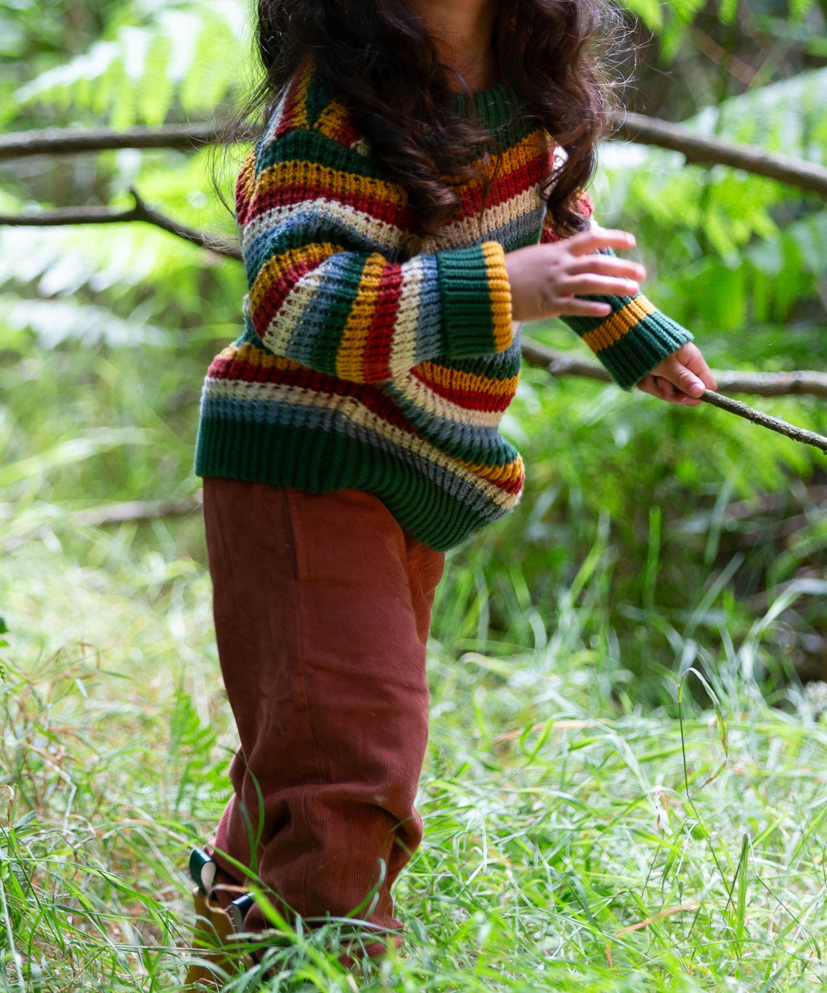 A close up of a child walking through woodland and wearing the Little Green Radicals rainbow waffle snuggly knitted jumper showing the waffle knit detail
